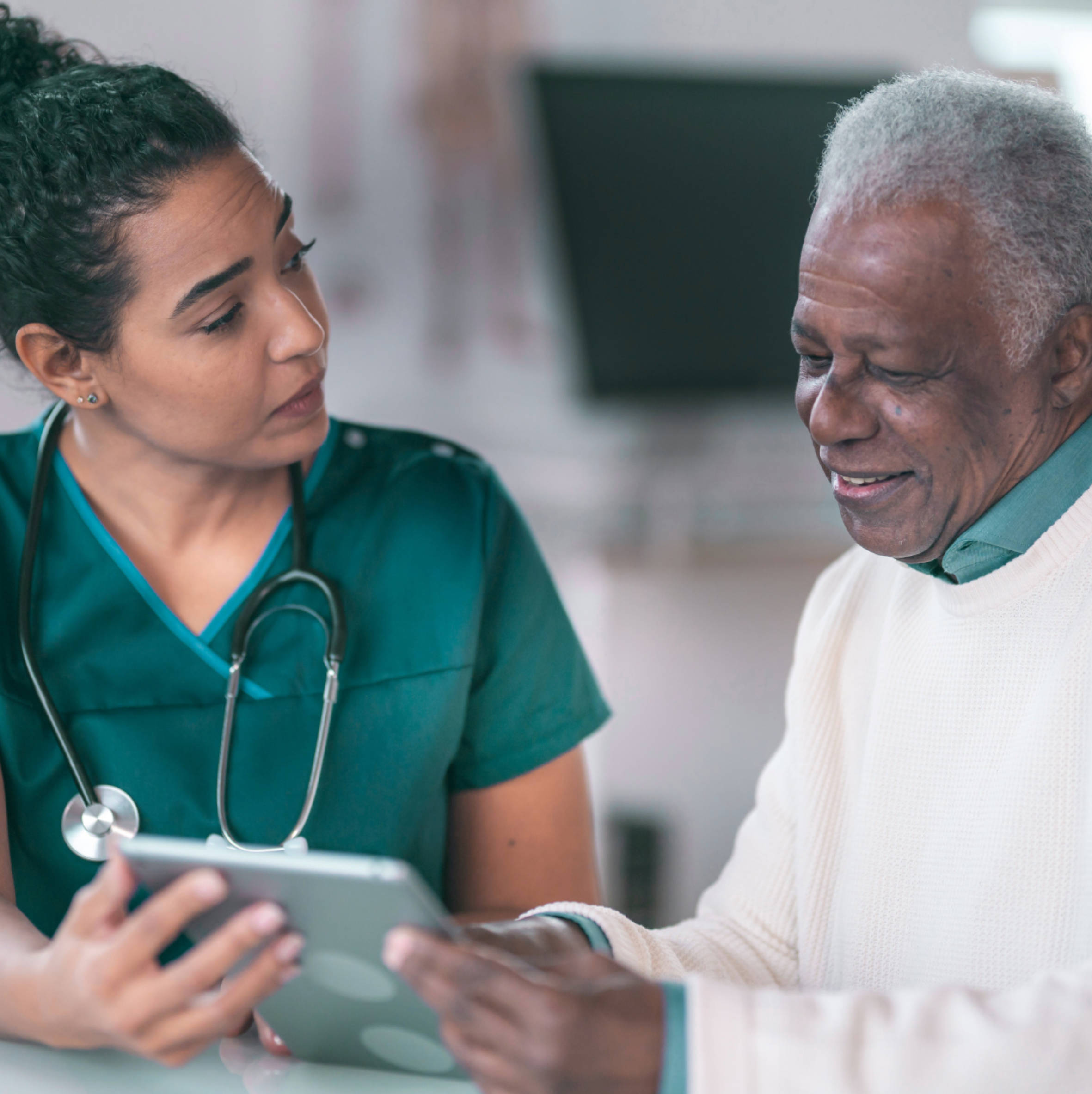 Female healthcare professional in scrubs showing a tablet to an elderly man who is smiling.